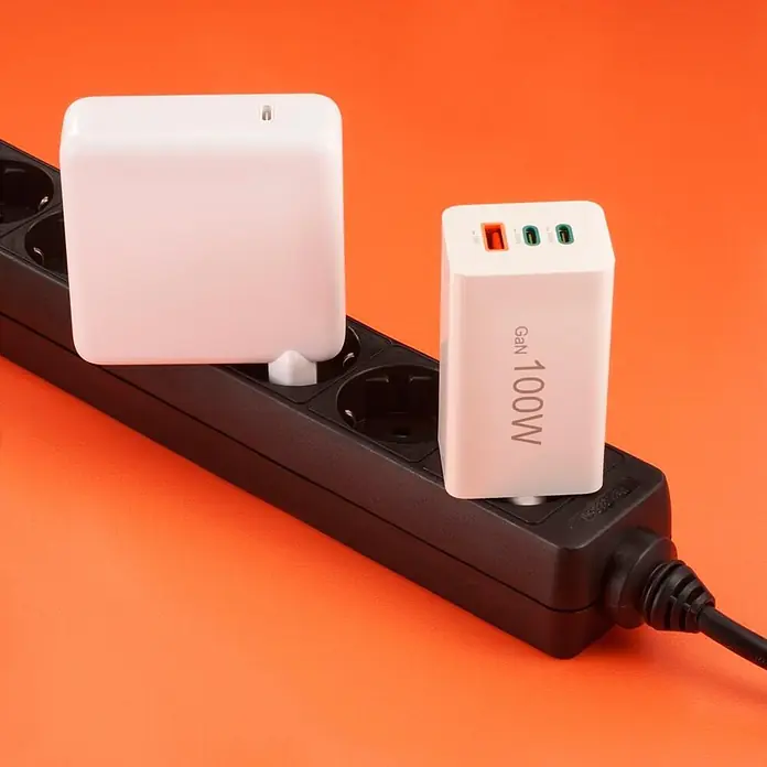Hama Schnellladegerät 00201997 GaN 2x USB-C 1x USB-A Mini-Ladegerät PD 100 W - Weiß - Zwei weiße Netzteile, eines GaN 100W, in einer schwarzen Steckerleiste auf orangefarbenem Untergrund eingesteckt.