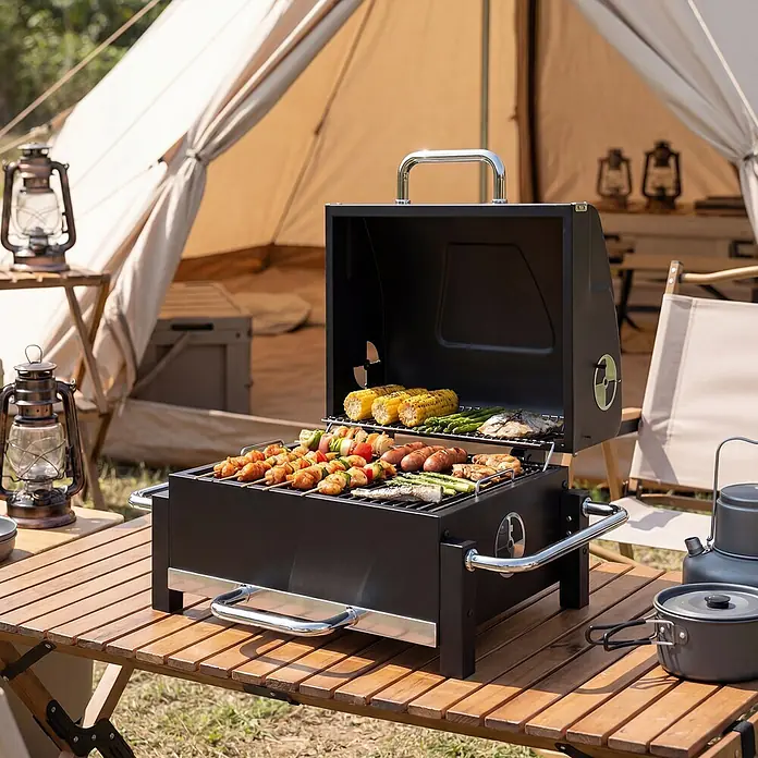 Outsunny Smoker -  - Ein schwarzer, tragbarer Grill mit Chromgriffen, auf einem Holztisch beim Camping. Er brät verschiedene Speisen.