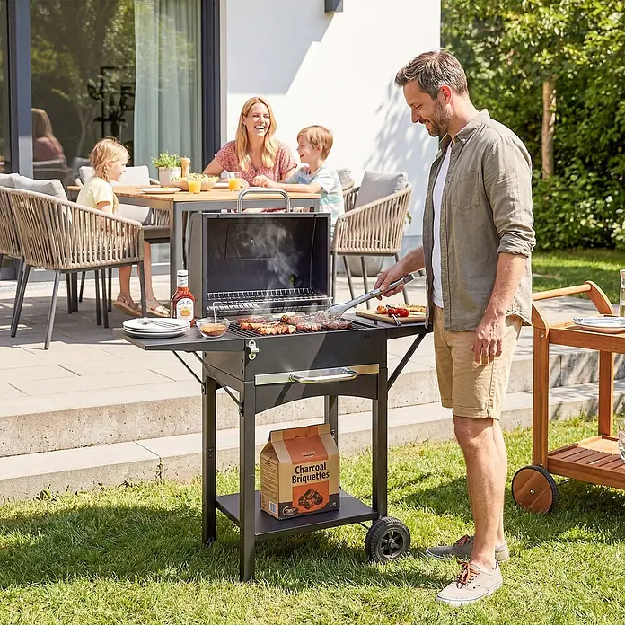 Outsunny Smoker -  - Schwarzer Holzkohlegrill mit Seitenablagen und Rollen, aktiv zum Grillen bei einem Familien-Barbecue im Freien genutzt.