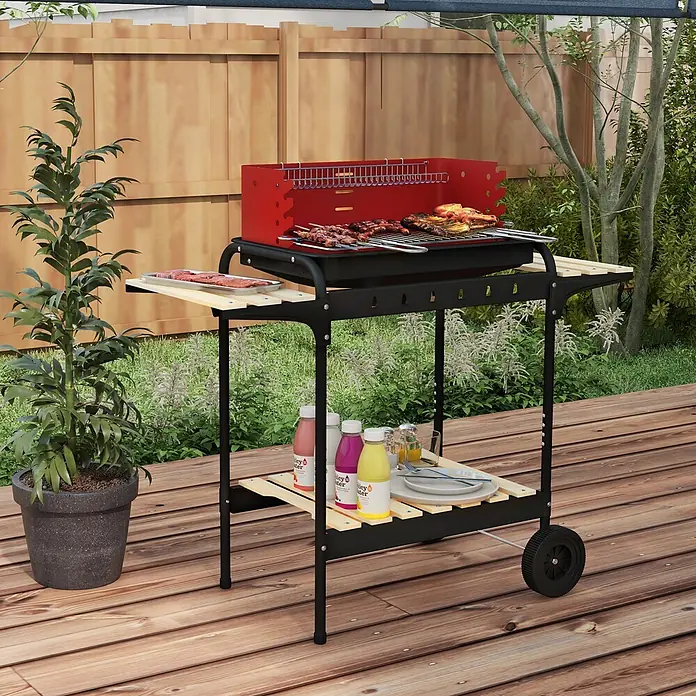 Outsunny Smoker -  - Ein rot-schwarzer Grill mit Holzseitenablagen, auf dem Spieße und Hähnchen garen, steht auf einer Holzterrasse im Garten.