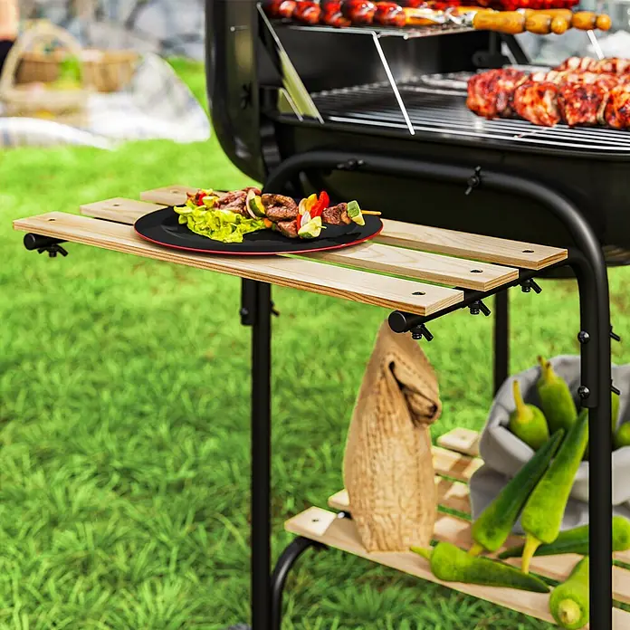 Outsunny Holzkohlegrill - Durchmesser Grillfläche: 46 cm, Schwarz - Schwarzer Grill mit hellen Holz-Seitenablage und unterem Regal, mit Speisen im Garten.