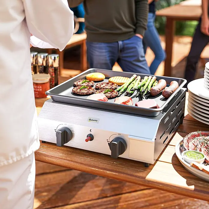 Outsunny Gasgrill - Anzahl Brenner: 2 Stk. - Silberner Tisch-Gasgrill mit schwarzer Grillplatte, der draußen auf einem Holztisch Burger, Spargel und Würstchen zubereitet, mit einem Koch in der Nähe.
