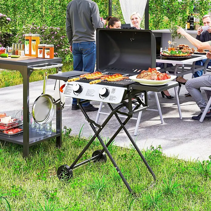 Outsunny Gasgrill - Anzahl Brenner: 2 Stk. - Ein schwarzer und weißer tragbarer Gasgrill mit Klappständer und Rädern, neben einem grauen Servierwagen, beim Grillen im Garten.