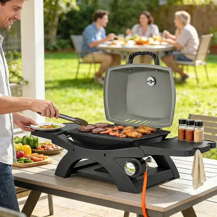 Outsunny Gasgrill - Anzahl Brenner: 1 Stk. - Ein Mann benutzt einen dunkelgrauen tragbaren Gasgrill, bereitet Burger, Garnelen und Hähnchenflügel im Freien zu, während andere im Hintergrund speisen. Grill auf Holztisch.