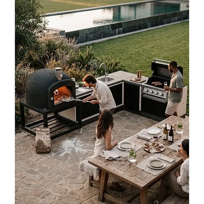 Tuozi Kitaway Outdoorküchen-Modul - Grau/Weiß, B x T x H: 100 x 100 x 65 cm - Schwarzer Outdoor-Pizzaofen und Küchenzeile mit Grill und Spüle, von Personen im Garten zum Kochen und Essen genutzt.