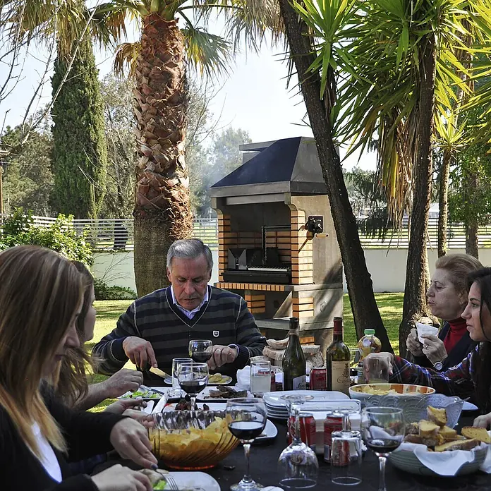 Movelar Pan American Holzkohlegrill - Schwarz/Orange - Familie genießt ein Grillessen im Freien mit einem gemauerten Grill unter Palmen.