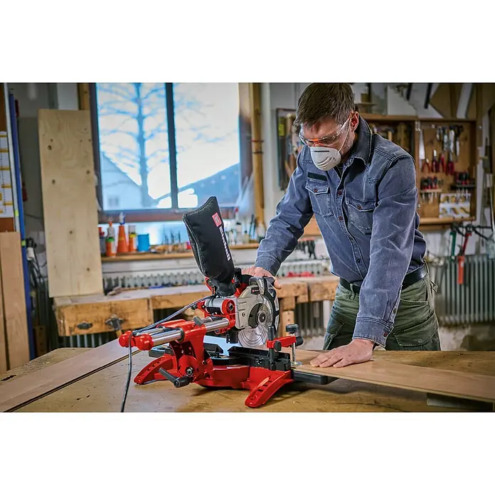 Einhell Kapp- & Gehrungssäge TC-SM 2131/1 Dual - 1.800 W, Durchmesser Sägeblatt: 210 mm, Leerlaufdrehzahl: 4.900 U/min - Mann in Schutzausrüstung bedient eine rot-silberne Kappsäge zum Holzzuschnitt.