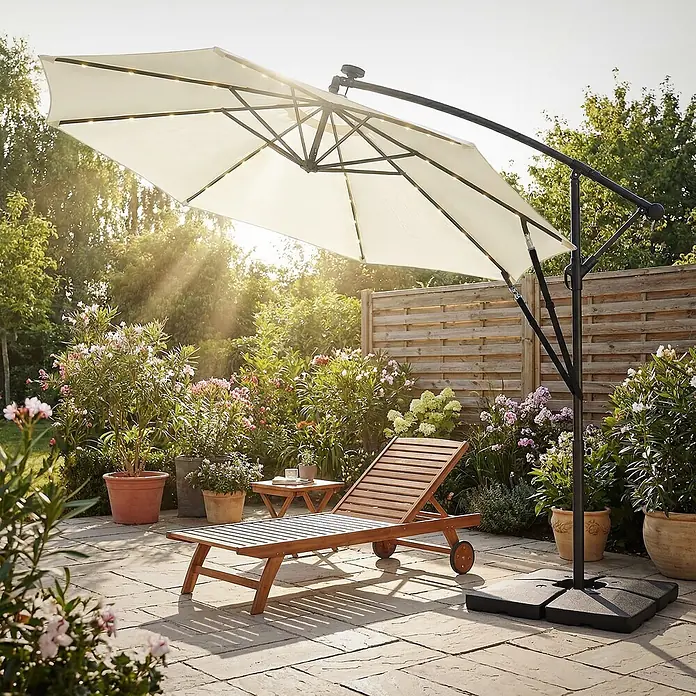 ML-Design Ampelschirm rund Gartenschirm UV50+ Sonnenschutz wasserabweisend 360° drehbar & neigbar - Beige, Durchmesser Bespannung: 300 cm - Großer weißer freitragender Gartenschirm mit integrierten Solarleuchten beschattet eine Holzliege und einen Tisch auf einer Terrasse.