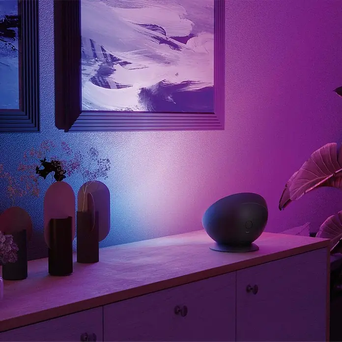 Hama Deko-Tischleuchte RGBW - Schwarz - Dunkles, ovales Smart-Gerät auf einem Holzschrank, beleuchtet von violettem und blauem Umgebungslicht.