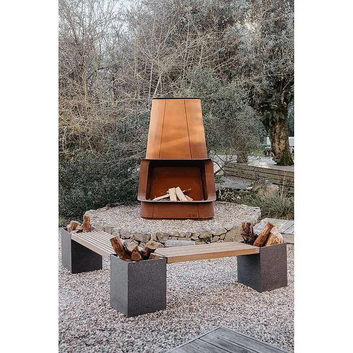 Tuozi Feuerschale Apollo - L x B x H: 125 x 53 x 190 cm, Cortenstahl - Rostbrauner Cortenstahl-Kamin mit Holzscheiten und passender Holzbank mit integriertem Holzlager auf einer Kiesterrasse.