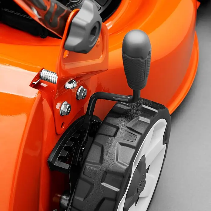 Husqvarna Benzin-Rasenmäher LB244E - 2.200 W, 139 cm³, Schnittbreite: 440 mm - Nahaufnahme des Rads und Höhenverstellhebels eines orangefarbenen Rasenmähers mit grauem Knopf und Metallfeder.