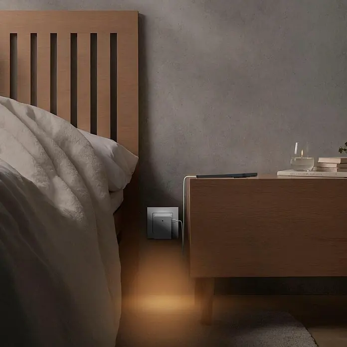 Hama LED-Nachtlicht 00223506 1x USB-A 1x USB-C Dämmerungssensor -  - Kleines graues Nachtlicht steckt in einer Wandsteckdose neben einem Bett und strahlt einen warmen Schein auf den Boden.