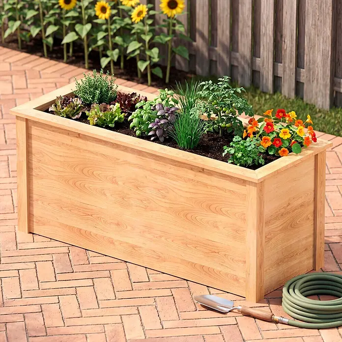 Dobar Hochbeet - 150 x 60 x 65 cm, Holz, Natur - Helles Holz-Hochbeet, gefüllt mit verschiedenen Kräutern, Salaten und Blumen, auf einer Ziegelterrasse mit Gartengeräten.