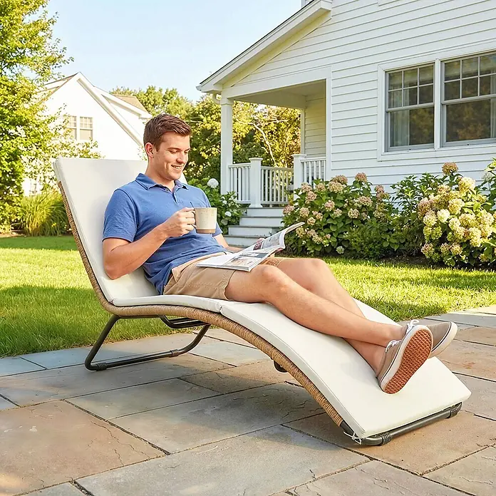 Outsunny Gartenliege - Cremeweiß, Stahl - Geflochtene Outdoor-Chaiselongue mit cremefarbenem Kissen, von einem Mann zum Entspannen und Lesen in einem sonnigen Garten genutzt.