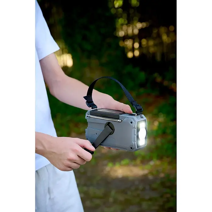 Thomson Radio de batería solar de emergencia RT280G - Gris, 8,8 x 16,8 x 7 cm, AM / FM / LINTERNA - Persona girando la manivela de una radio de emergencia gris con linterna integrada, panel solar y correa negra, al aire libre con la luz encendida.