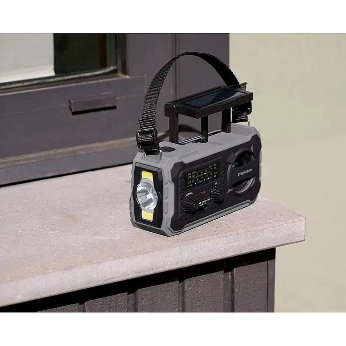 Thomson Radio de batería solar de emergencia RT280G - Gris, 8,8 x 16,8 x 7 cm, AM / FM / LINTERNA - Una radio de emergencia portátil gris y negra con panel solar integrado y linterna, sobre un alféizar de piedra al aire libre.
