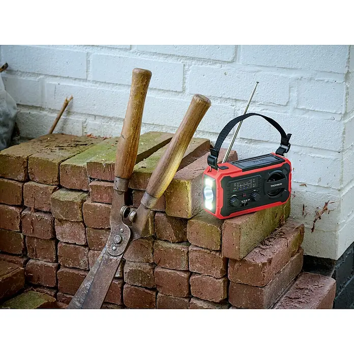 Thomson Radio de batería solar de emergencia RT280R - Rojo, 8,8 x 16,8 x 7 cm, AM / FM / LINTERNA - Radio Thomson roja y negra con linterna encendida y antena, junto a tijeras de podar sobre ladrillos.