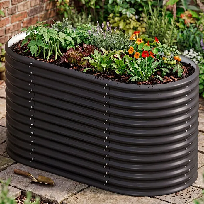 Vitavia Hochbeet Terra 858 - 162 x 82 x 85,8 cm, Zincalume®, Schwarz - Dunkelgraues gewelltes ovales Hochbeet, gefüllt mit verschiedenen Pflanzen, mit einer kleinen Schaufel daneben.
