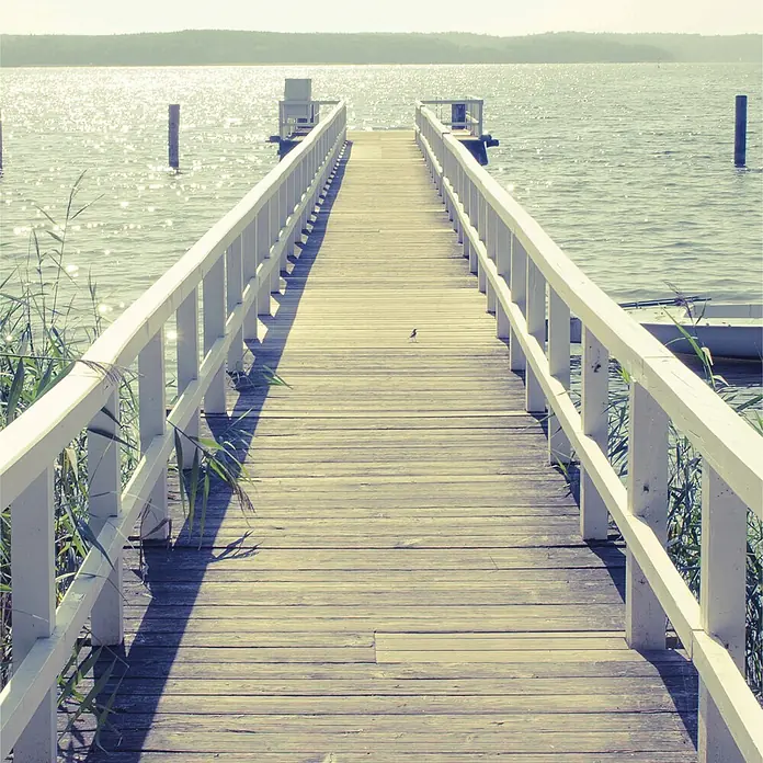 SanDesign Alu-Verbundplatte - 100 x 250 cm, Jetty into Sea - Langer Holzsteg mit weißen Geländern erstreckt sich über sonnenbeschienenes Wasser. Ferne Küstenlinie sichtbar.