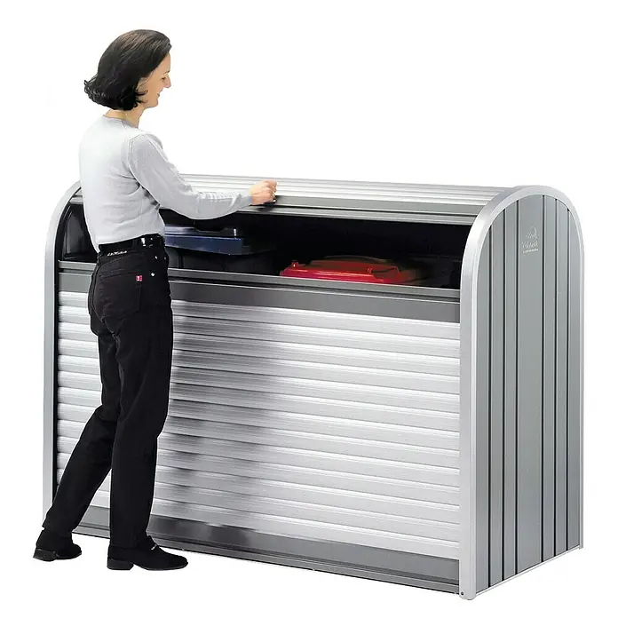 Biohort StoreMax Garten-Aufbewahrungsbox StoreMax 190 - Quarzgrau, 190 x 97 x 136 cm, Stahl - Frau öffnet eine Mülltonnenbox aus Metall mit Rollladen, in der blaue und rote Mülltonnen stehen.\n
