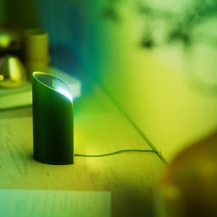 Philips Hue LED-Tischleuchte Play Wallwasher - L x B x H: 9,1 x 7,6 x 15,7 cm, Weiß, RGBW - Grüne zylindrische Schreibtischlampe, die helles grün-gelbes Umgebungslicht auf einem Holztisch abstrahlt.