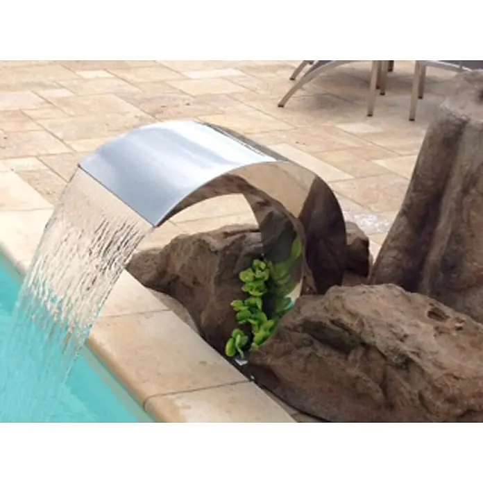 Ideal Eichenwald Pooldusche Modell 52 Schwalldusche Nevada - Edelstahl - Glänzender Edelstahl-Wasserfallauslauf, der sich in einen Pool wölbt, umgeben von Steinen und Grün.