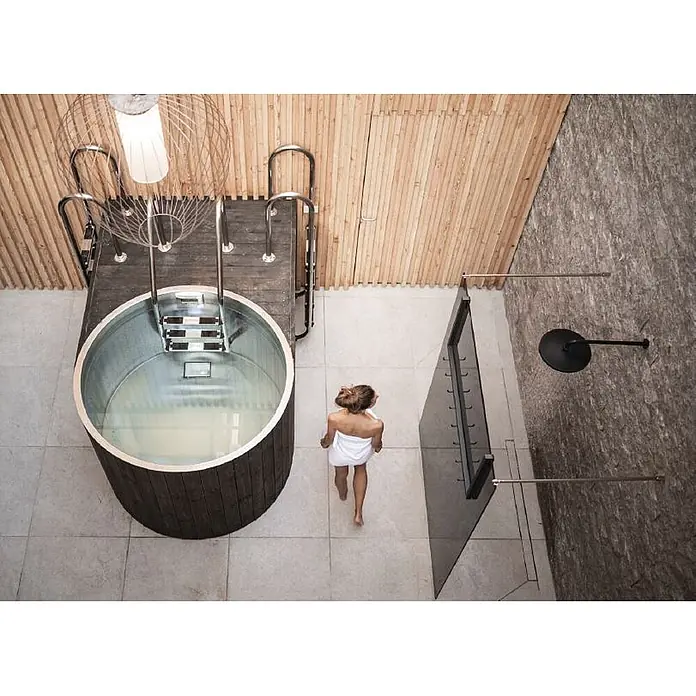 Ideal Eichenwald Pooldusche Modell 35 Gartendusche Nemo mit Piezo Taster - Edelstahl - Person nähert sich einem runden Tauchbecken aus dunklem Holz und Metall mit Stufen, neben einer modernen Dusche im Wellnessbereich.