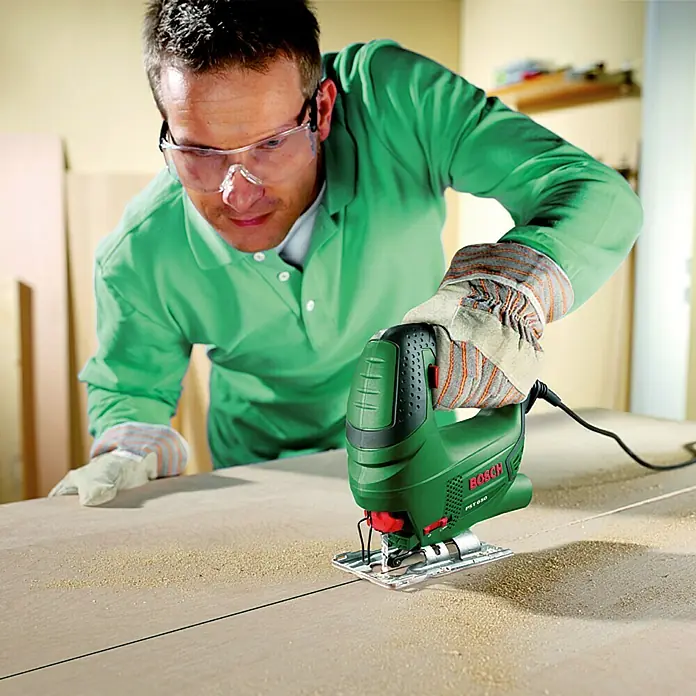Bosch Elektro-Stichsäge PST 650 - 3.100 /min, 65 mm - Ein Mann sägt ein Holzbrett mit einer grünen Bosch Stichsäge, wobei er Schutzbrille und Handschuhe trägt. Das Werkzeug gleitet präzise über das Holz.