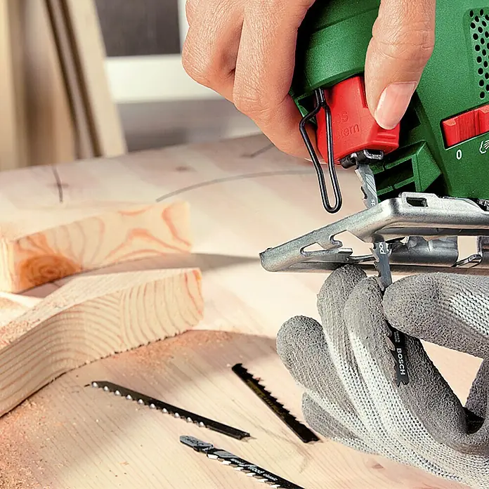 Bosch Elektro-Stichsäge PST 650 - 3.100 /min, 65 mm - Eine Nahaufnahme einer Stichsäge, bei der ein Sägeblatt eingesetzt wird, mit Holzstücken im Hintergrund.
