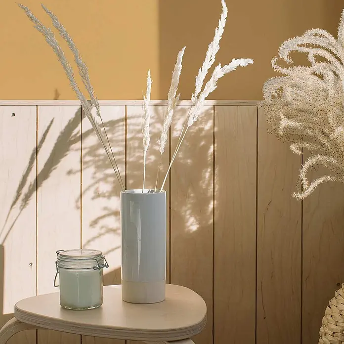 Tadessi Wandfarbe Bernstein - Gelb, Matt, 2,5 l - Weiße Keramikvase mit getrocknetem weißem Gras, daneben eine Kerze im Glas auf einem Holztisch.