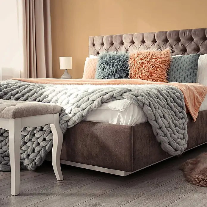 Tadessi Wandfarbe Kamelbraun - Beige, Matt, 5 l - Ein gemütliches Bett mit gestepptem Kopfteil, dunklem Rahmen, grob gestrickter Decke, pfirsichfarbenem Überwurf und strukturierten Kissen in einem warmen Schlafzimmer.