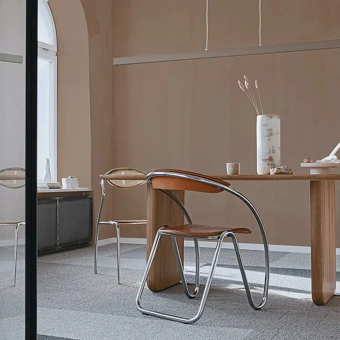 Tadessi Wandfarbe Oliv - Beige, Matt, 1 l - Moderner Stuhl mit brauner Ledersitzfläche und Chromgestell, an einem hellen Holztisch platziert.