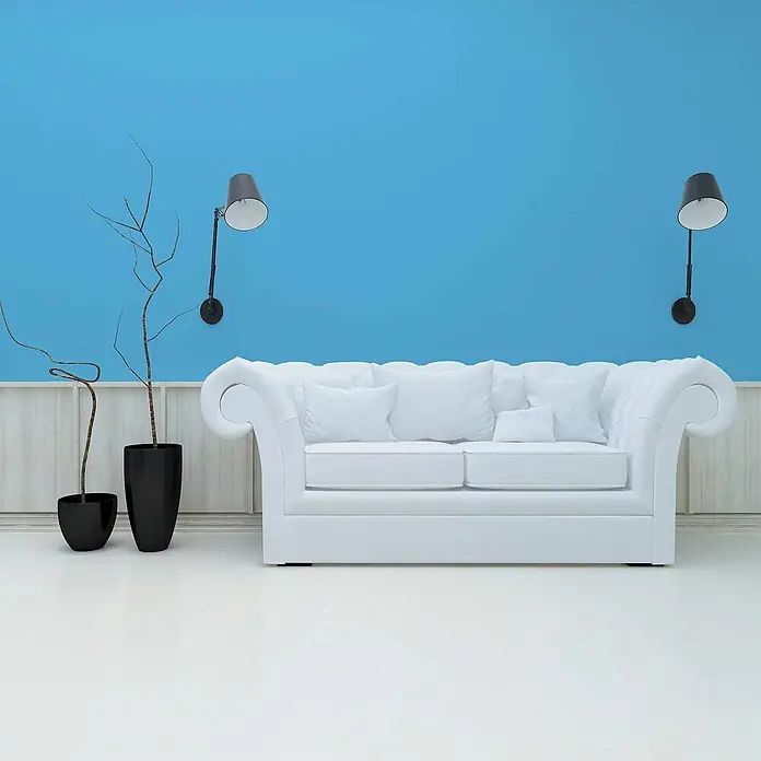 Tadessi Wandfarbe Azurblau - Blau, Matt, 2,5 l - Weißes, getuftetes Sofa mit Rollarmen in einem Raum mit blauer Wand, schwarzen Wandlampen und Zierzweigen.