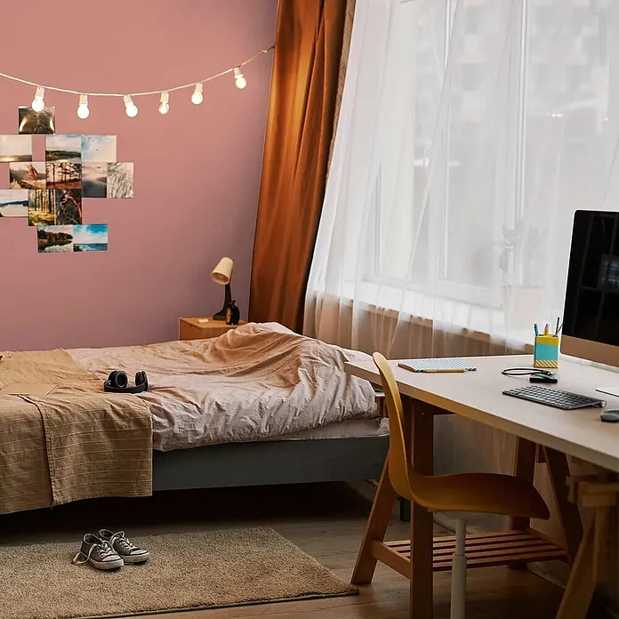 Tadessi Wandfarbe Rosenholz - Rot, Matt, 1 l - Modernes Schlafzimmer mit Bett, hellem Holzschreibtisch, Computer und Lichterkette an rosafarbener Wand.