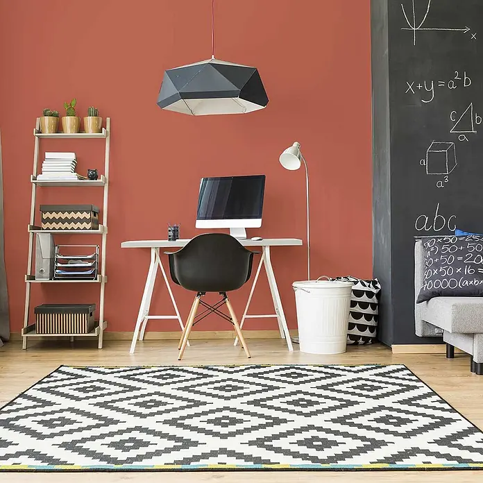 Tadessi Wandfarbe Honig - Rot, Matt, 2,5 l - Ein modernes Heimbüro mit roter Wand, weißem Schreibtisch, schwarzem Stuhl und einem großen schwarz-weißen Geometrie-Teppich.