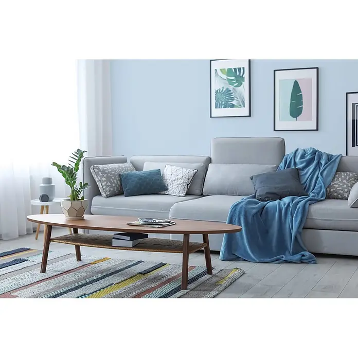 Tadessi Wandfarbe Himmelblau - Blau, Matt, 2,5 l - Ovaler Holztisch mit geflochtener Ablage vor einem grauen Sofa, mit Pflanze und Büchern.