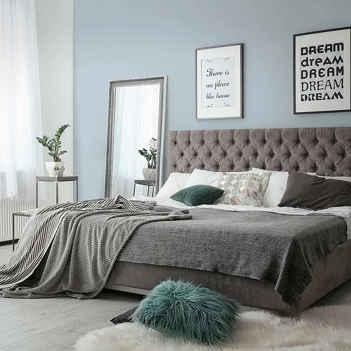 Tadessi Wandfarbe Pastellblau - Blau, Matt, 1 l - Ein geräumiges, graues Polsterbett mit getuftetem Kopfteil, mit Decken und Kissen in einem hellblauen Schlafzimmer.