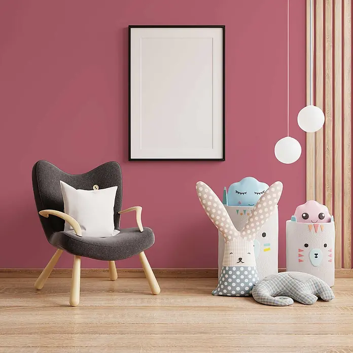 Tadessi Wandfarbe Eisrosa - Pink, Matt, 5 l - Ein vertikaler schwarzer Bilderrahmen mit weißem Passepartout an einer rosa Wand, neben einem grauen Sessel und Spielzeugkörben im Kinderzimmer.