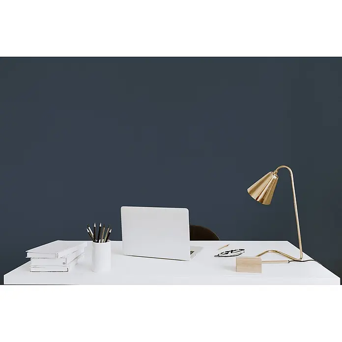 Tadessi Wandfarbe Schieferblau - Blau, Matt, 5 l - Minimalistisches Home Office mit weißem Schreibtisch, silbernem Laptop, goldener Schreibtischlampe und Büromaterial vor dunkelblauer Wand.