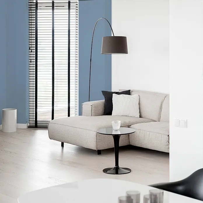 Tadessi Wandfarbe Eisblau - Blau, Matt, 2,5 l - Modernes Wohnzimmer mit hellgrauem L-Sofa, dunkler Stehlampe, hellem Holzboden und blau-weißen Wänden.