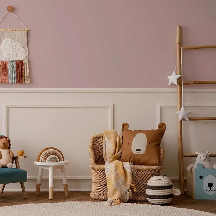 Tadessi Wandfarbe Altrosa - Rosa, Matt, 1 l - Ein modernes Kinderzimmer mit rosa Wänden, weißer Täfelung, einem gewebten Wolken-Wandteppich und natürlichen Rattanmöbeln.