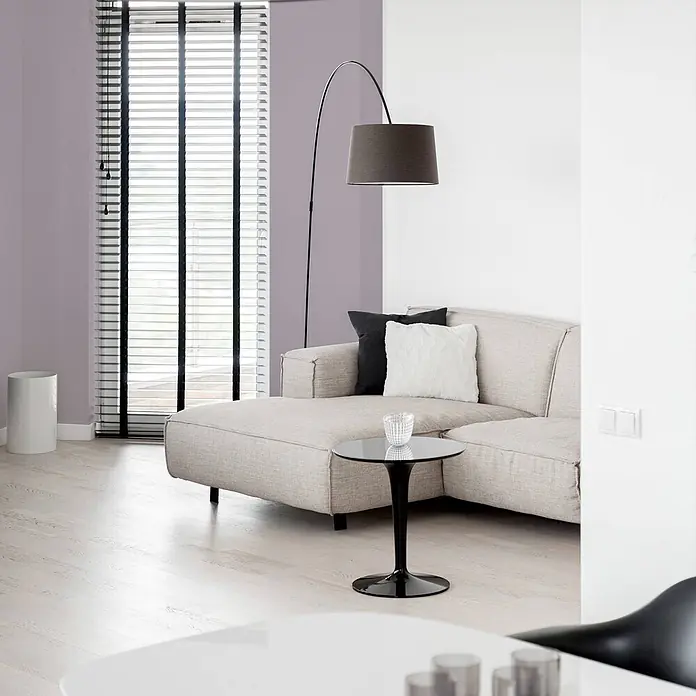 Tadessi Wandfarbe Lavendel - Violett, Matt, 1 l - Modernes Wohnzimmer mit beigem Ecksofa, dunkler Bogen-Stehlampe und kleinem schwarzem Beistelltisch. Heller Boden und lila Akzentwand.