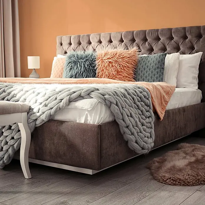 Tadessi Wandfarbe Kupfer - Orange, Matt, 5 l - Ein gemütliches Bett mit dunkelgrauem, getuftetem Kopfteil, bezogen mit farbigen Kissen und einer grob gestrickten Decke in einem warmen Schlafzimmer.