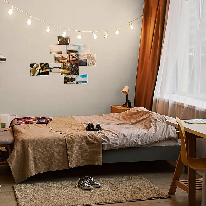 Tadessi Wandfarbe Karamell - Beige, Matt, 1 l - Gemütliches Schlafzimmer mit einem Bett mit hellbrauner Bettwäsche und Lichterkette mit Fotos, das eine warme, einladende Atmosphäre schafft.