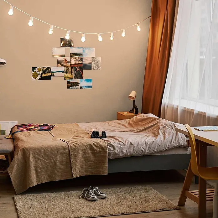Tadessi Wandfarbe Honigbeige - Beige, Matt, 5 l - Gemütliches Schlafzimmer mit einem Bett, beiger Decke, Lichterkette und Fotocollage an einer hellbraunen Wand, in warmes Licht getaucht.