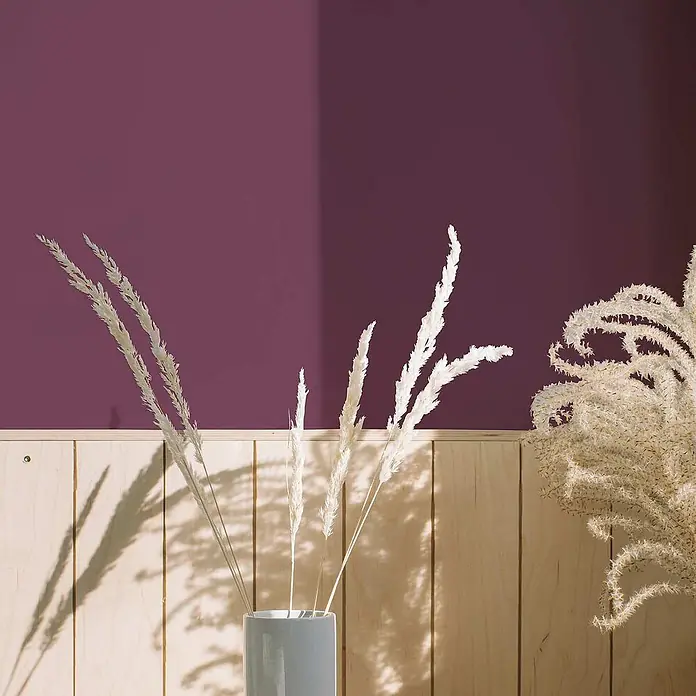 Tadessi Wandfarbe Aubergine - Violett, Matt, 1 l - Getrocknete weiße Ziergräser in einer grauen Vase, ausgestellt auf Holzpaneelen vor einer lila Wand.
