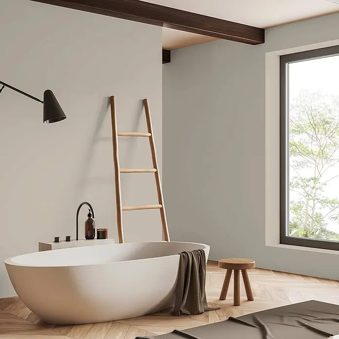 Tadessi Wandfarbe Sandgrün - Beige, Matt, 1 l - Eine hellbeige freistehende Badewanne in einem modernen Badezimmer, mit einem dunklen Handtuch am Rand. Steht auf Holzboden neben einem Fenster.