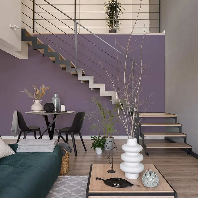 Tadessi Wandfarbe Magenta - Violett, Matt, 1 l - Modernes Wohnzimmer mit dunkelgrünem Sofa, Holztisch und Essbereich vor einer lila Akzentwand und offener Treppe.