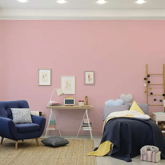 Tadessi Wandfarbe Sandrosa - Pink, Matt, 5 l - Ein gemütliches pinkes Zimmer mit blauem Sessel, Holzschreibtisch und Bett mit blauer und gelber Bettwäsche. Gerahmte Bilder an der Wand.