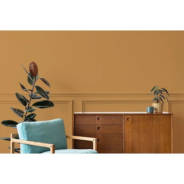 Tadessi Wandfarbe Ocker - Orange, Matt, 5 l - Senfgelbe Wand mit Paneelen, hellblauer Sessel, Holzkommode und zwei Topfpflanzen.